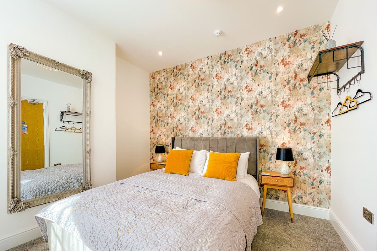 A bedroom with a large mirror, floral wallpaper, a bed with a quilt, two bedside tables with lamps, and a wooden headboard.