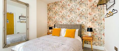 A bedroom with a large mirror, floral wallpaper, a bed with a quilt, two bedside tables with lamps, and a wooden headboard.
