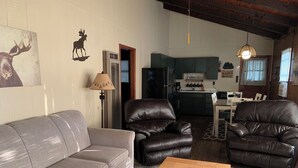 Una sala de estar con un sofá gris, dos sillones de cuero, una mesa de centro de madera y una cocina al fondo.