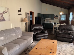 A living room with a grey sofa, two leather armchairs, a wooden coffee table, and a kitchen in the background.