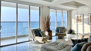 Una sala moderna con un amplio ventanal corredera que da a un balcón con vistas al mar. Hay dos sillones y un sofá con cojines.