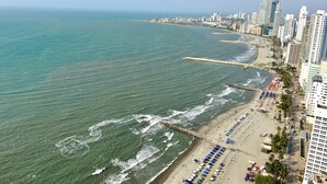 Un paisaje urbano costero con una playa bordeada por edificios altos, un muelle largo que se adentra en el mar y numerosas sombrillas de playa.