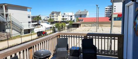A deck with wicker chairs and a barbecue grill, overlooking a beach and buildings.