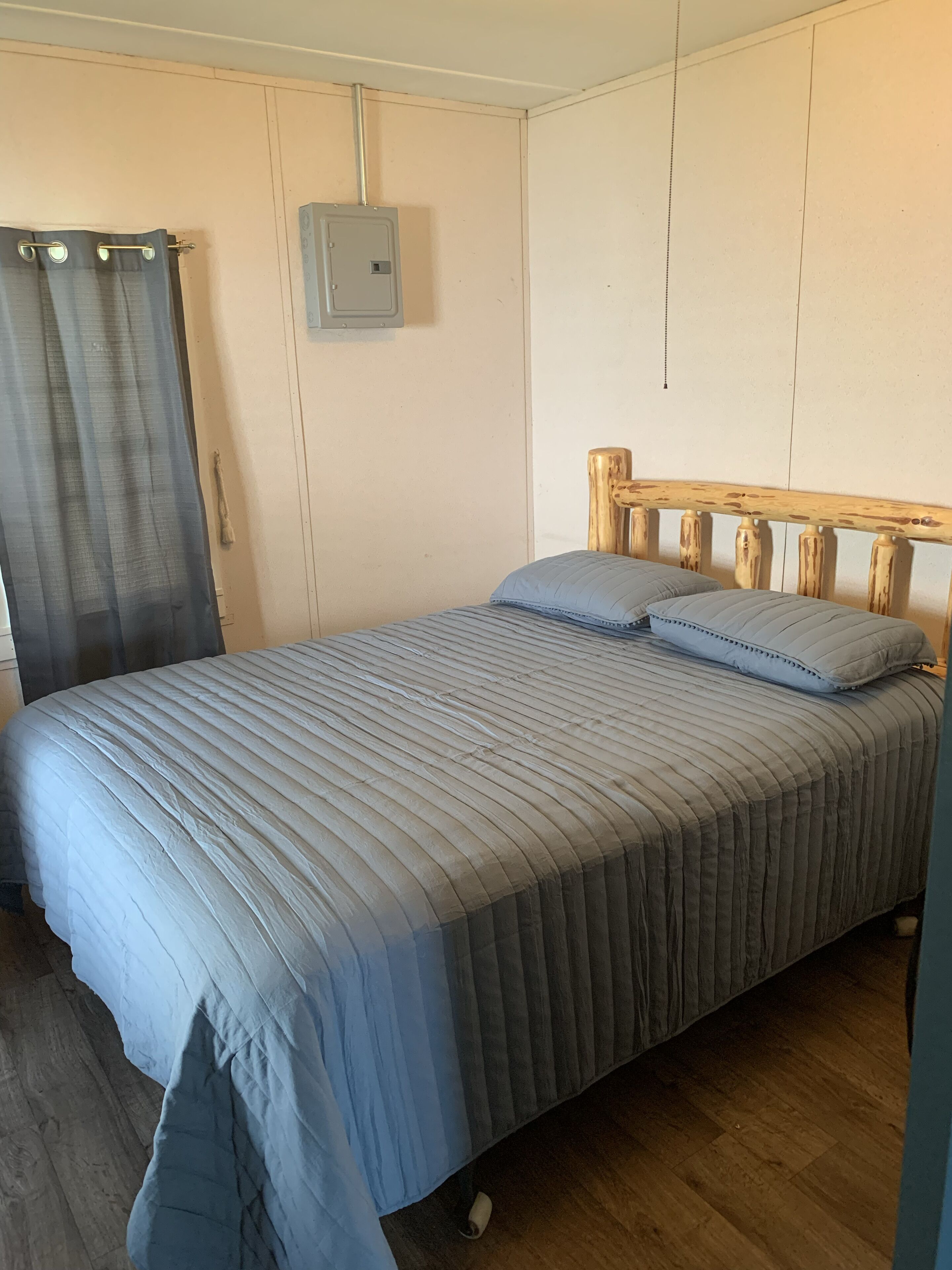 A single bed with a wooden headboard, a grey bedspread, and two pillows. A grey curtain is partially drawn, and a light switch is mounted on the wall above the bed.