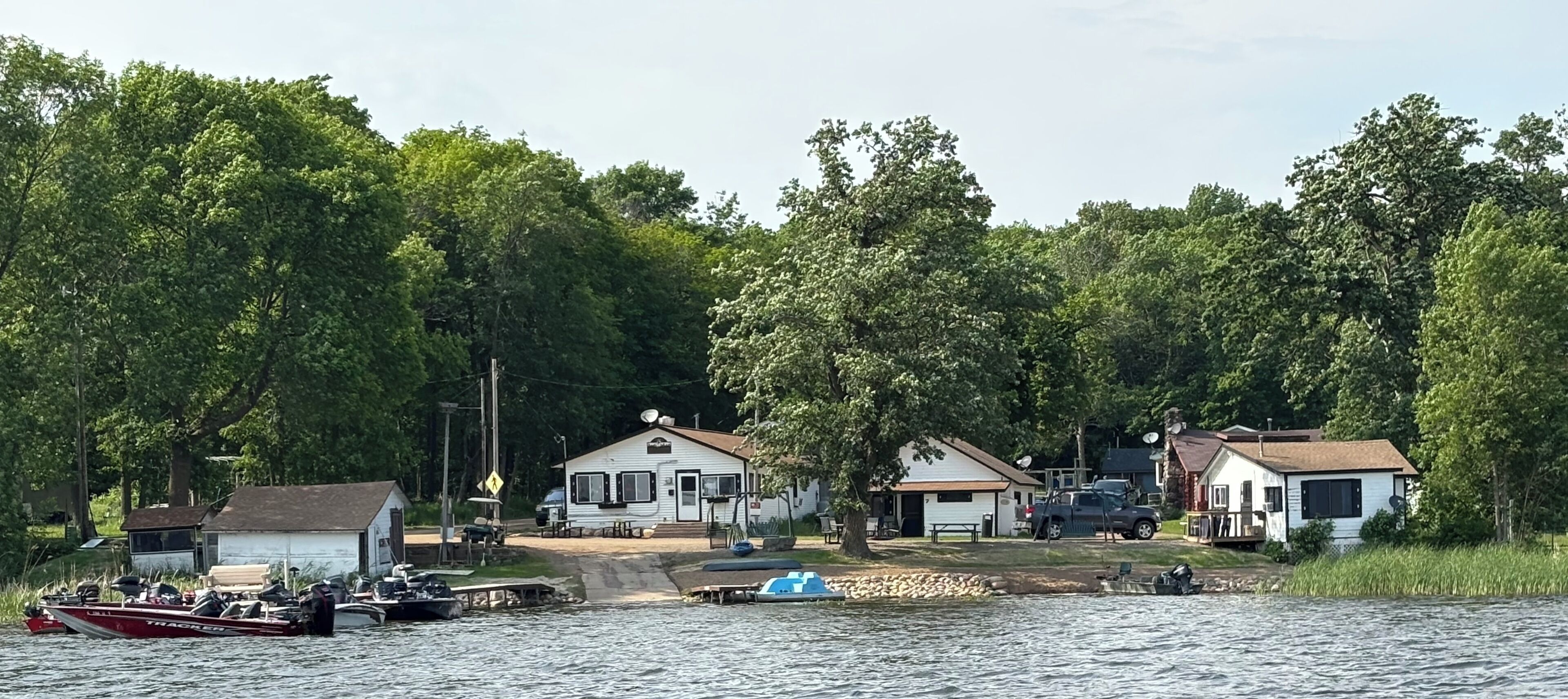 Une propriété au bord du lac comprenant plusieurs chalets, un quai avec des bateaux et un petit bateau amarré.