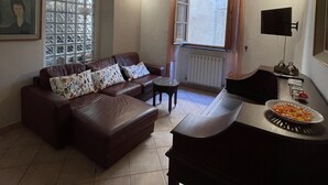 A living room with a brown leather sofa, a small round table, a piano, and a window with curtains. - Artemisia Pisa (Pisa)