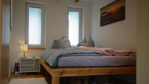 Een slaapkamer met een houten bedframe, twee ramen met zonwering, een schilderij aan de muur en een witte nachtkastje met een lamp.