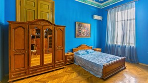 A bedroom with a large wooden wardrobe, a bed with a blue bedspread, a painting on the wall, and a window with curtains.