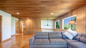 Ein geräumiges Wohnzimmer mit Holzboden, einem grauen Ecksofa, einem Deckenventilator und Blick durch große Fenster.