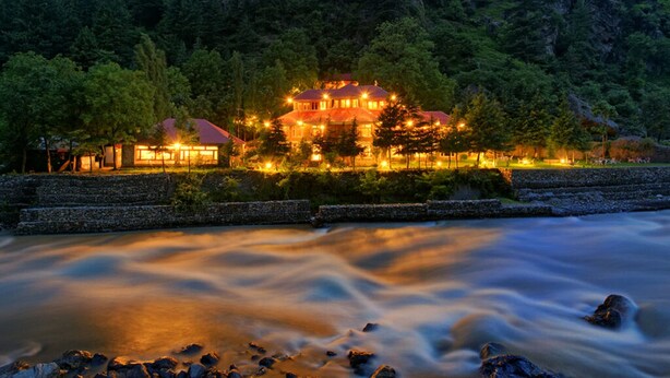 A riverside hotel with a stone embankment and a forested hillside. - Pine Park Edge Resort Khanian (Balakot)