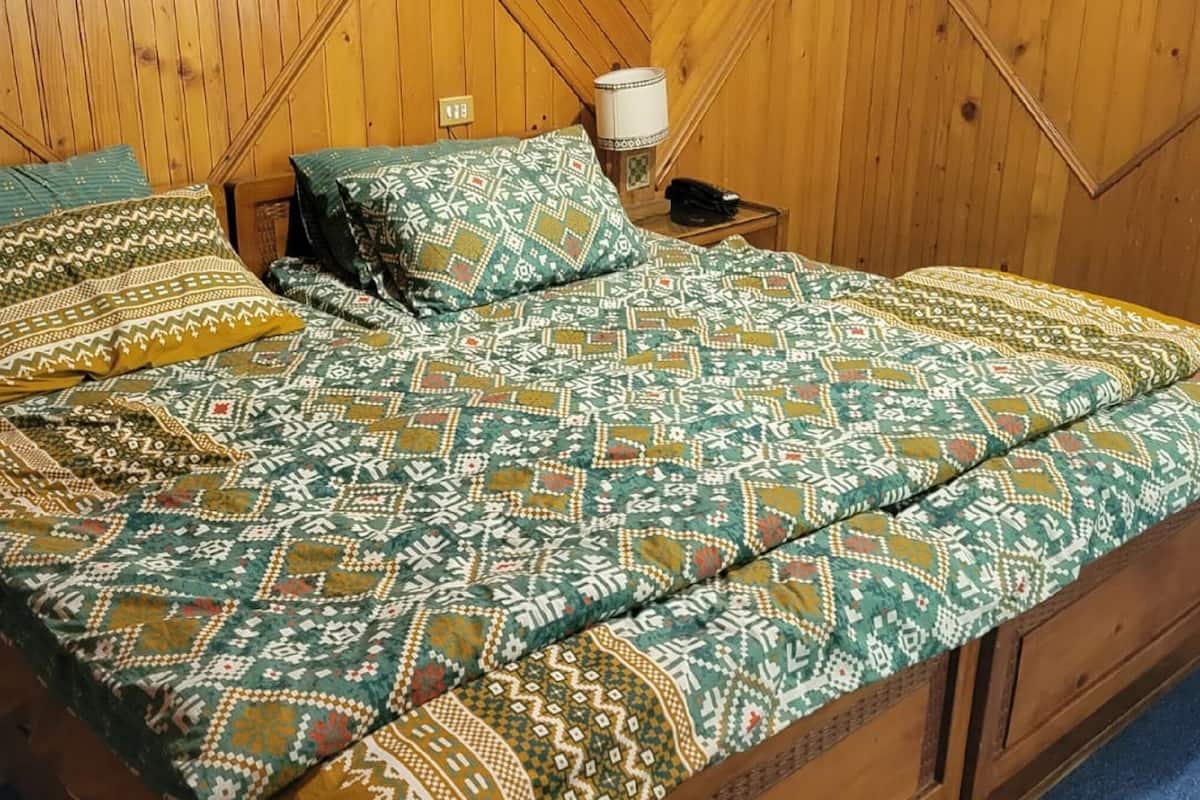 Ein Zimmer mit Holzwänden, einem Bett mit gemusterten Textilien, einer Nachttischlampe und einem Heizgerät.