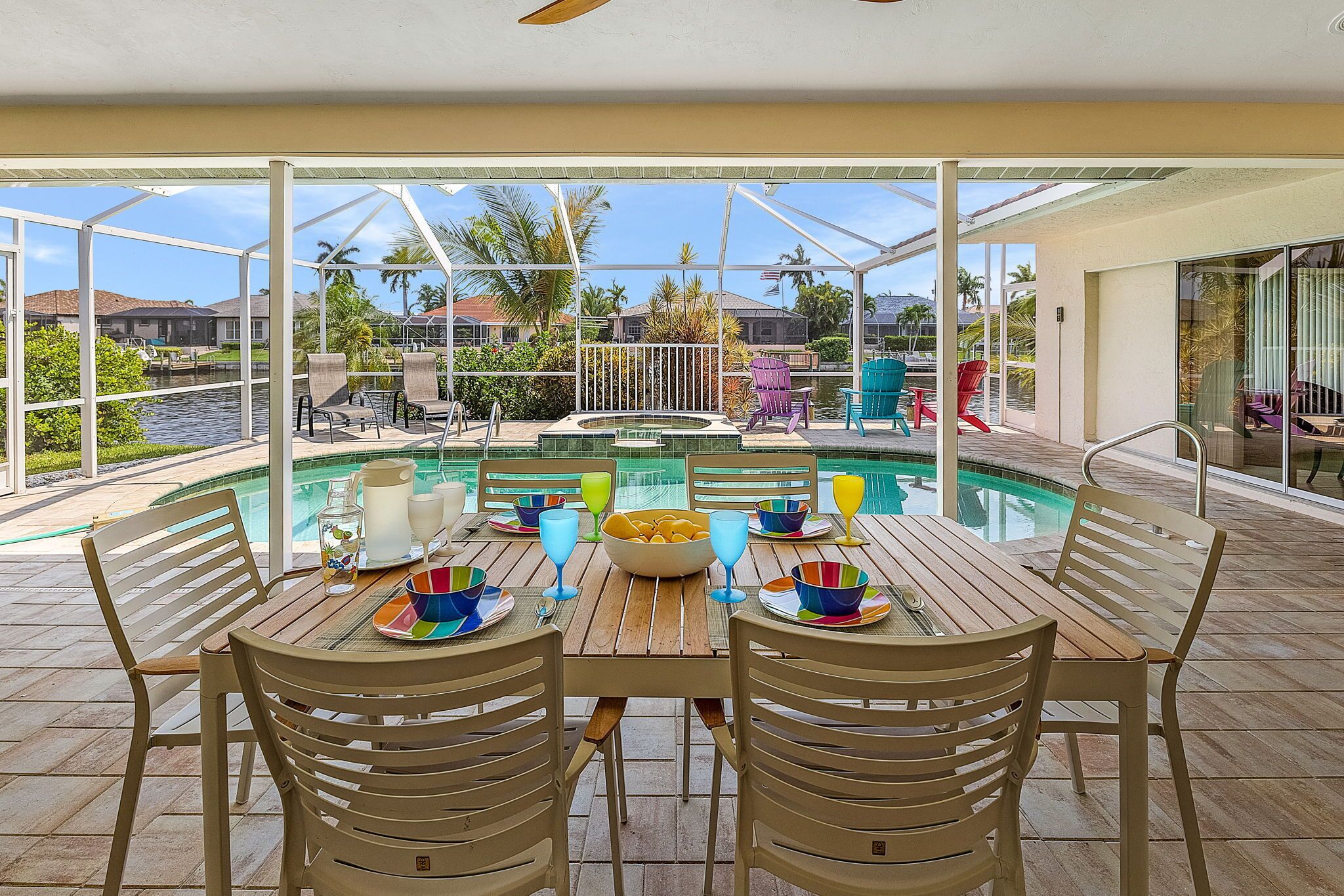 An outdoor dining area with a pool, wooden tables, and chairs, surrounded by glass walls and palm trees.