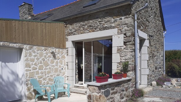 Une maison en pierre avec une terrasse en bois, deux chaises turquoise et des plantes en pot sur le porche.