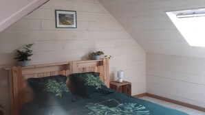 Une chambre avec un plafond en pente, une tête de lit en bois, un lit recouvert d’une couverture verte, deux tables de chevet avec des lampes et un tableau encadré au mur.
