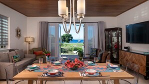 A modern dining room with a large table, clear glass chairs, and a view of the sea.