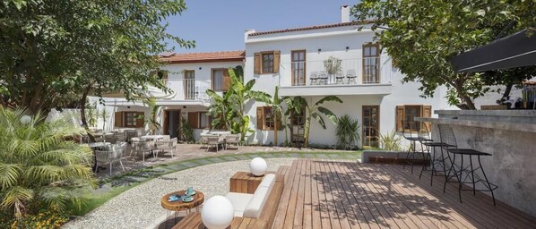 A two-story white building with a red-tiled roof, outdoor seating area with tables and chairs, and a wooden deck. - Mellow Blue Hotel (Seferihisar)