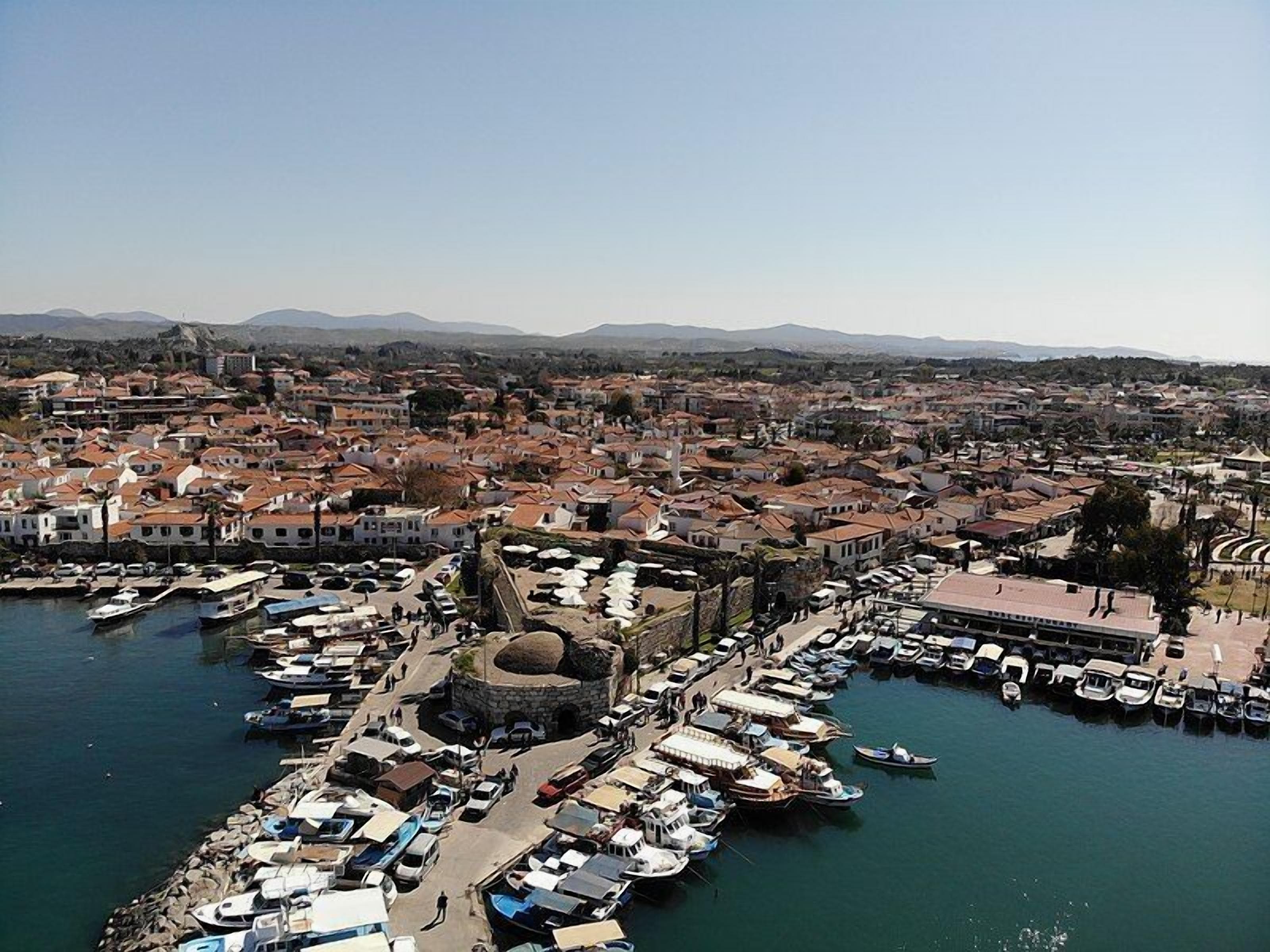 A coastal town with a marina filled with boats, a historic stone tower, and a dense urban area with red-roofed buildings.