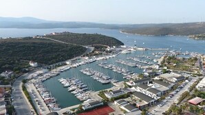 Vista aérea de un puerto deportivo con numerosas embarcaciones amarradas, rodeado de edificios y un paisaje montañoso al fondo.