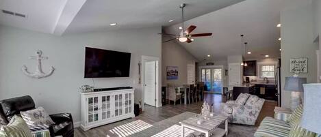 A spacious living room with a large TV, a ceiling fan, and a dining area with a kitchen in the background.