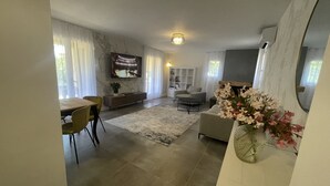 A modern living room with a dining area, a flat-screen TV mounted on the wall, and a large window with sheer curtains. - Private villa with pool (Saint-Raphaël)