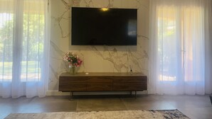A living room with a flat-screen TV mounted on a marble wall, a wooden console table with a vase of flowers, and sheer curtains on the windows. - Private villa with pool (Saint-Raphaël)