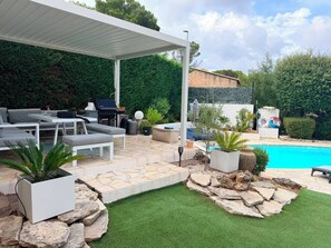 A covered outdoor seating area with a barbecue grill, a pool, and a well-maintained garden with artificial grass and potted plants. - Private Villa with Pool (Saint-Raphaël)