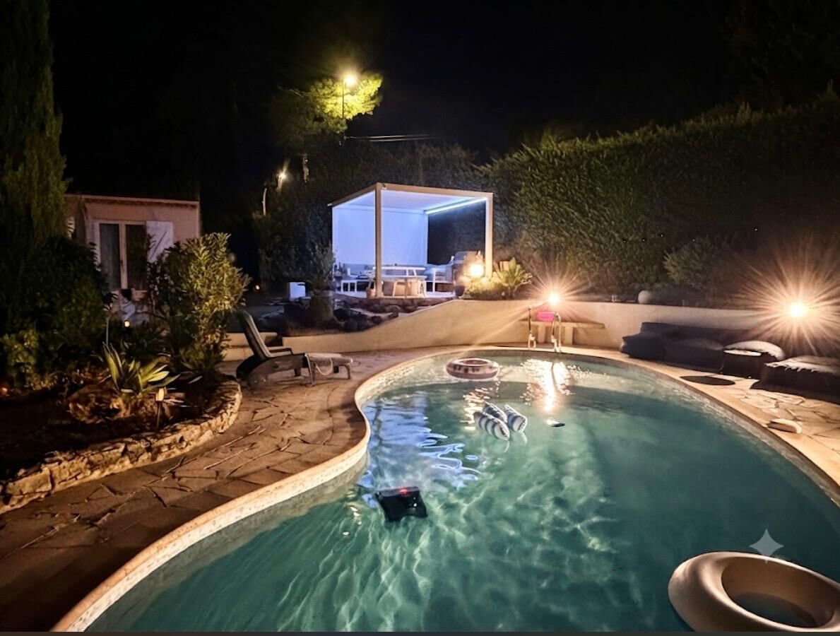 A swimming pool with a curved edge, surrounded by a stone deck. There are lounge chairs and a small covered area with a table and chairs.