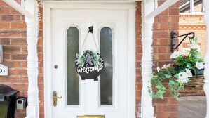 Eine weiße Haustür mit einem 'welcome'-Schild, einem Briefkasten und einem Hängetopf mit Blumen.
