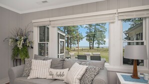 A cozy living room with a grey sofa, a small table with a lamp, and a view of a grassy area and trees through large windows. - Sea Glass Beach House!  Large sand beach, dock and hot tub! Pets welcome!  (Hague)