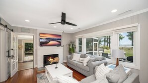A spacious living room with a fireplace, a large window with a view, and a ceiling fan. - Sea Glass Beach House!  Large sand beach, dock and hot tub! Pets welcome!  (Hague)