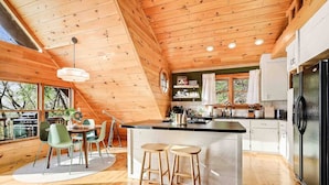 A modern kitchen with wooden beams, a central island, bar stools, and a dining area with chairs. - Modern Mountaintop A-Frame | Wineries, Views, Lake (Morganton)