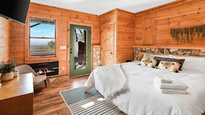 A wooden cabin bedroom with a bed, a fireplace, a view through a window, and a stone wall. - Modern Mountaintop A-Frame | Wineries, Views, Lake (Morganton)
