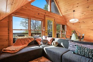 L’intérieur d’un chalet accueillant, avec un plafond en bois, de grandes fenêtres, un canapé avec des coussins et une table entourée de chaises.