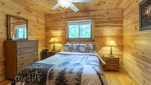 Une chambre à coucher dans une cabane en bois, avec un lit, deux lampes de chevet, une commode, un ventilateur de plafond et une fenêtre.
