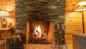 Une cheminée en pierre avec un feu, des murs en bois, une table avec une lampe et une chaise.