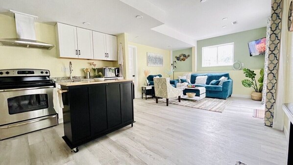 A modern kitchen with a stove, cabinets, and a black island. There is a living area with a blue sofa and a TV. - Cheerfull 2 bedroom Basement Apartment (Aurora)