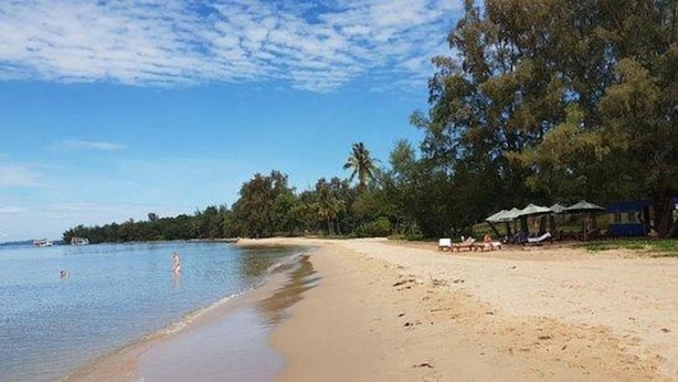 A beach with clear water, a sandy shore, and some trees. There are umbrellas and lounge chairs on the beach.