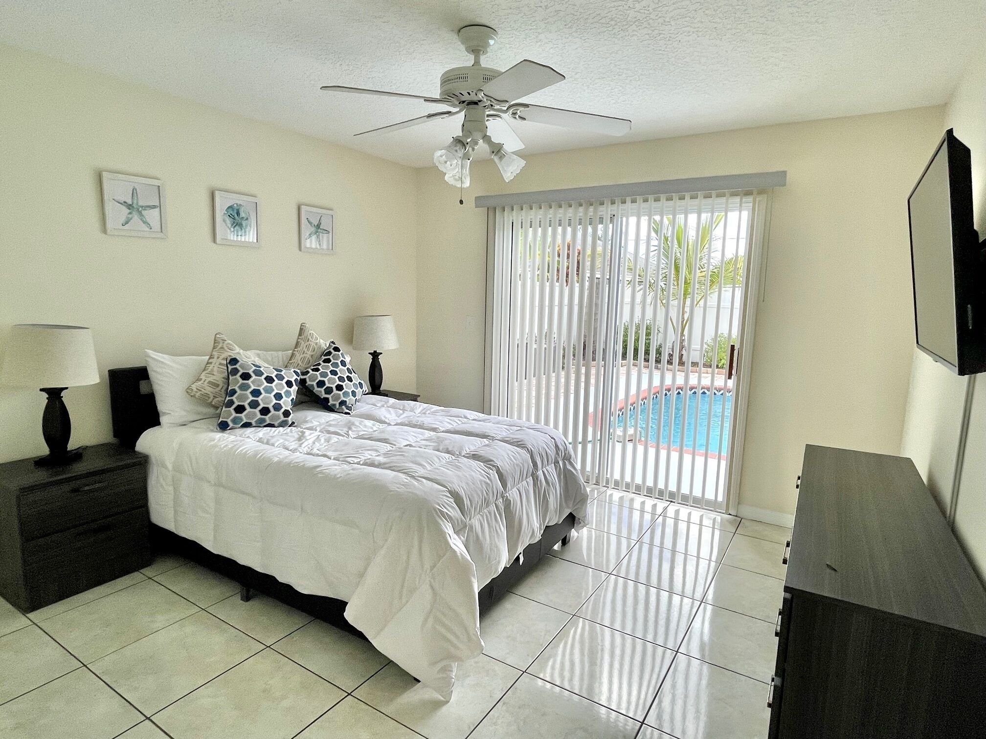 A bedroom with a bed, two bedside tables, a ceiling fan, a TV, and a window with blinds.