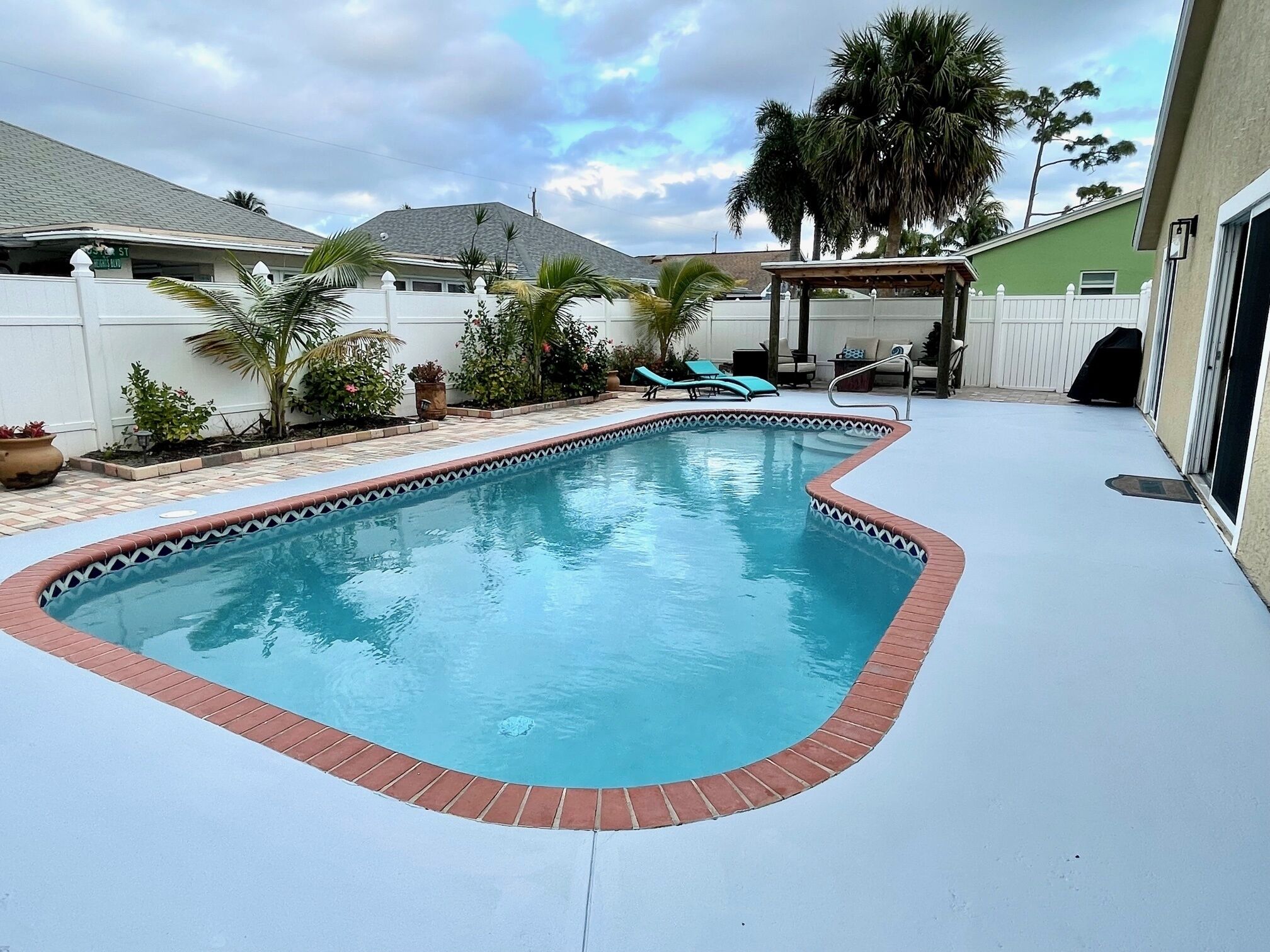 A rectangular swimming pool with a brick border, surrounded by a patio with a pergola and lounge chairs.