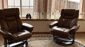 Deux fauteuils inclinables en cuir, placés face à une fenêtre donnant sur une terrasse en bois et un plan d’eau.