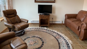 Un salon avec deux canapés en cuir marron, un grand téléviseur à écran plat posé sur un meuble en bois, un tapis à motifs et deux appliques murales.