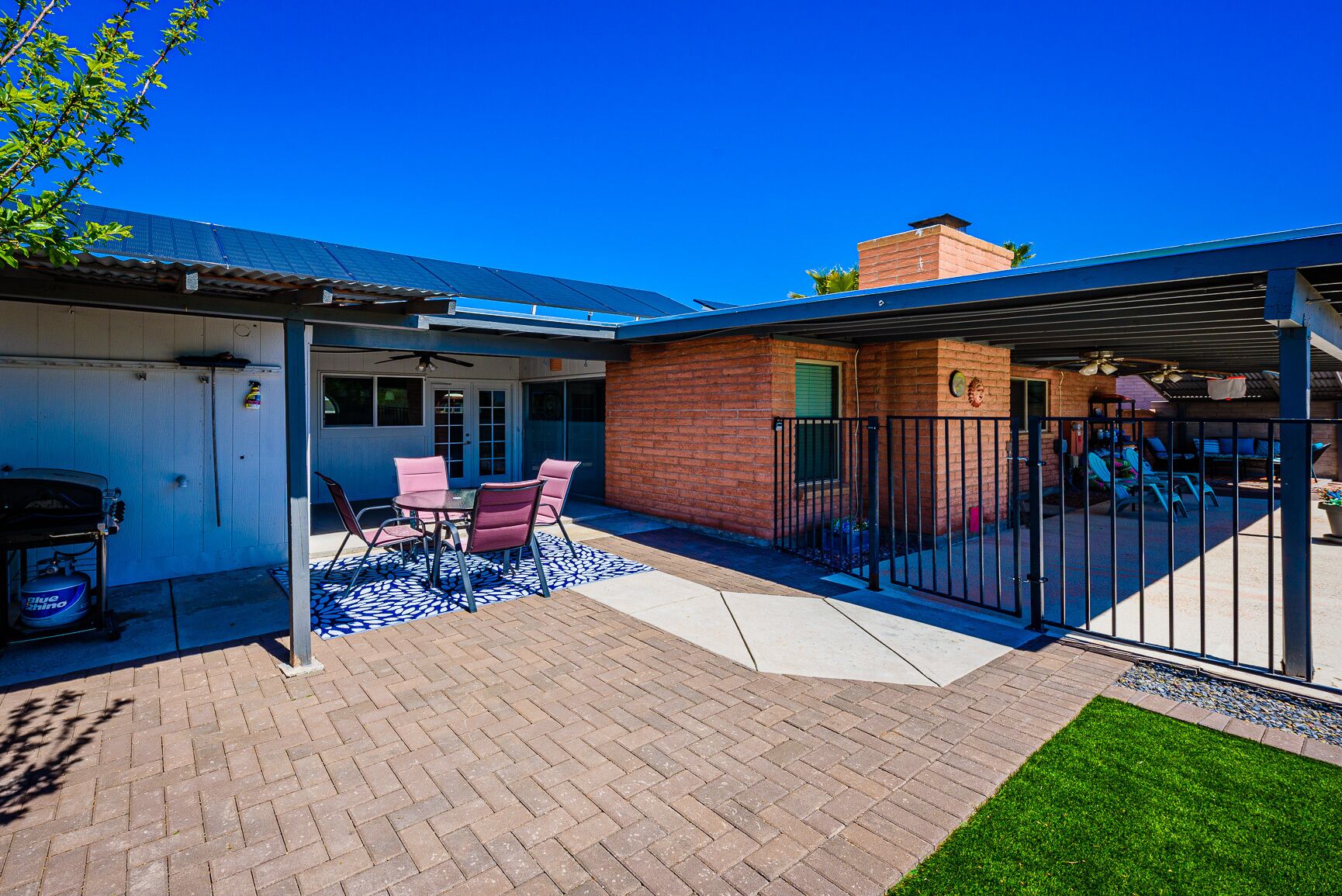 A patio with a brick floor, a covered seating area with a table and chairs, and a fenced backyard with a grill and outdoor furniture.