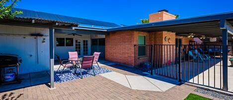 A patio with a brick floor, a covered seating area with a table and chairs, and a fenced backyard with a grill and outdoor furniture.