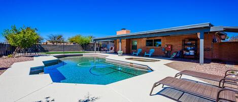 A rectangular swimming pool with a clear blue water feature, surrounded by a concrete deck. There are lounge chairs and a covered patio area with a brick wall.
