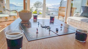 A table with a board game, two glasses of dark liquid, a bowl of snacks, and a view of the outdoors.