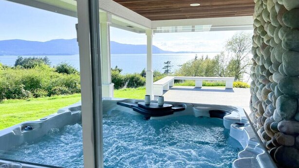 A hot tub with a view of mountains and a lake, surrounded by a stone wall and a wooden ceiling.