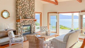 A cozy living room with a stone fireplace, wooden beams, and a view of the sea.