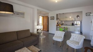 A living room with a sofa, two armchairs, a coffee table, and a lamp. There is a kitchen in the background with cabinets and a dining area. - Casa Chjara Maria (Vescovato)