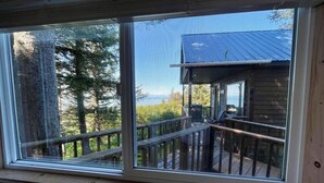 A room with a large window overlooking a wooden deck and a building with a blue roof. - Ocean-view Cabin on the Anchor Point Bluff (Anchor Point)