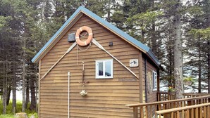 A wooden cabin with a sloped roof, a lifebuoy hanging on the exterior, a small window, and a welcome sign. - Ocean-view Cabin on the Anchor Point Bluff (Anchor Point)
